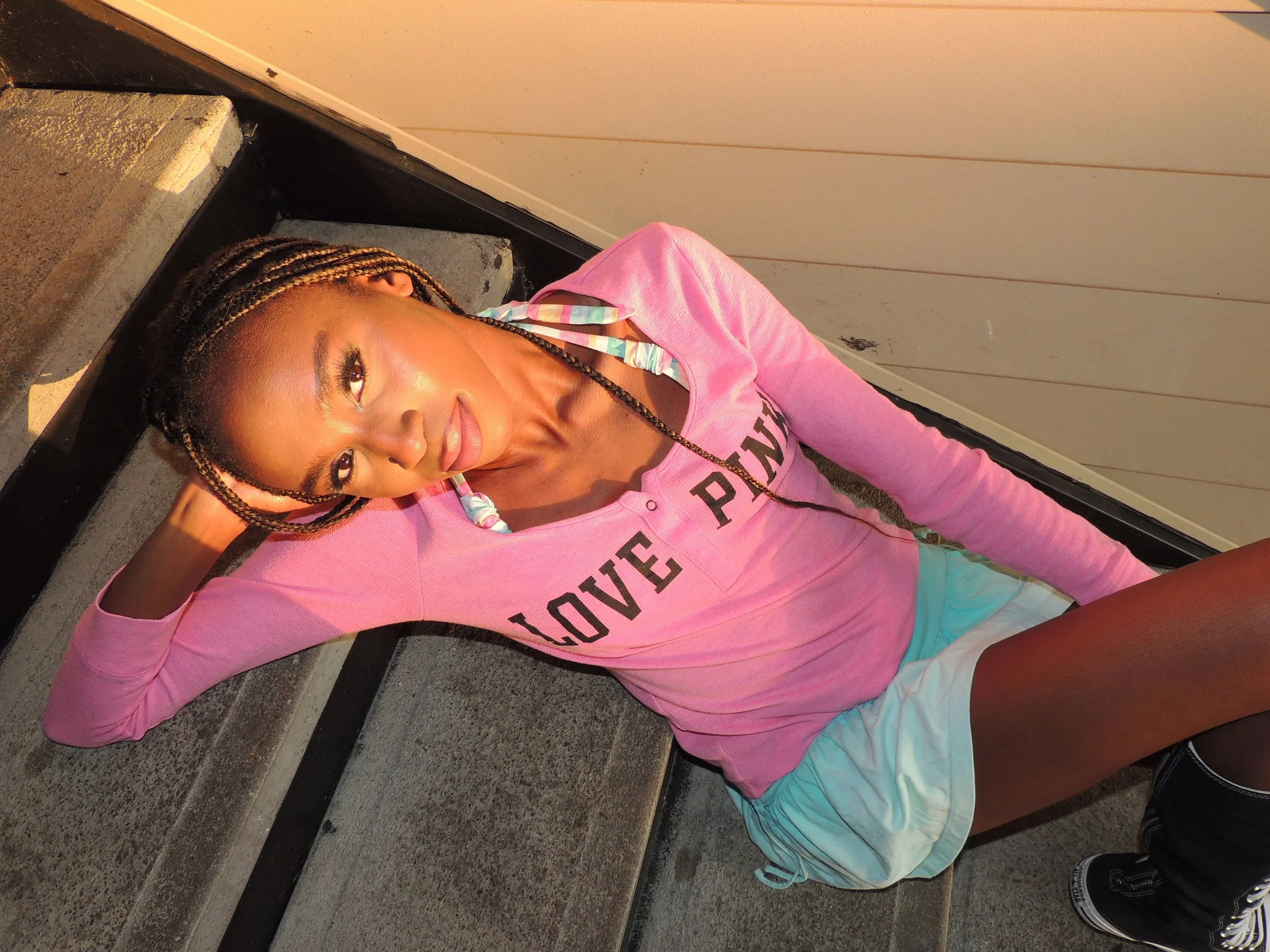 Young woman with braided hair and light skin lying on outdoor stairs, wearing a pink long-sleeve shirt with black letters and colorful trim, with a relaxed pose and sunlight on her face.