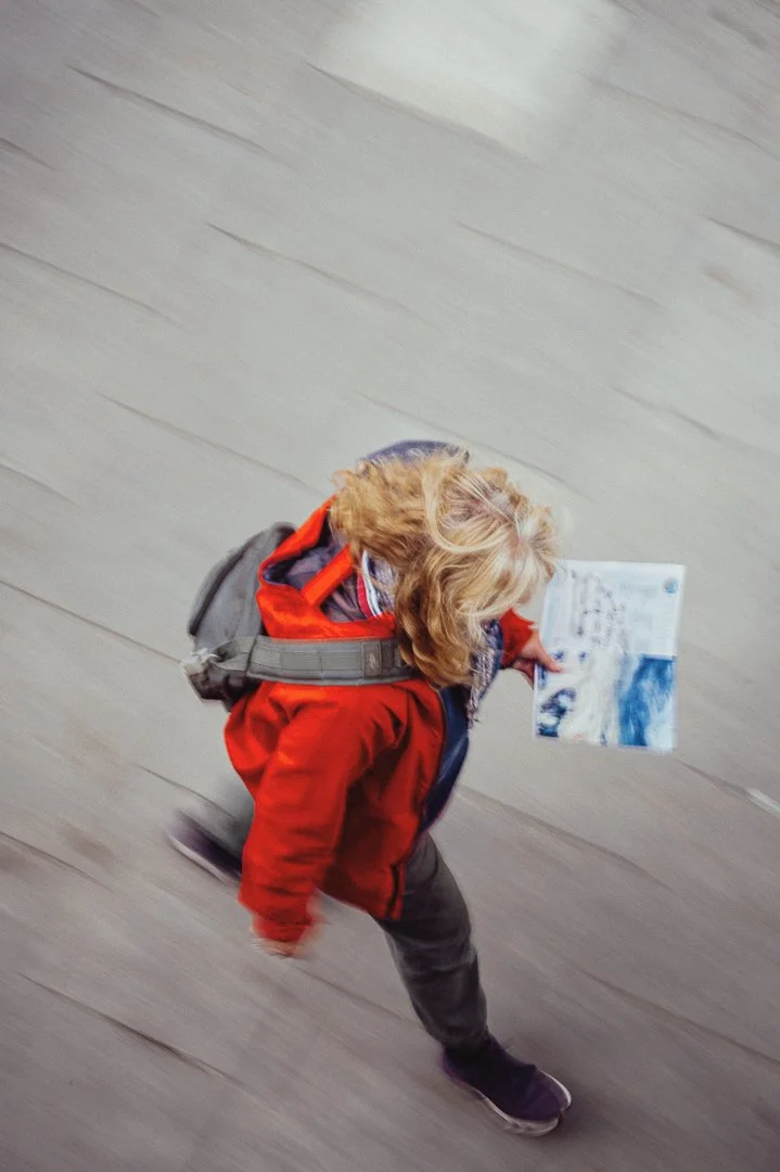 A young girl with blonde hair, wearing a red jacket and carrying a gray backpack, walking on a gray wooden floor while holding a booklet or small book with images and text.