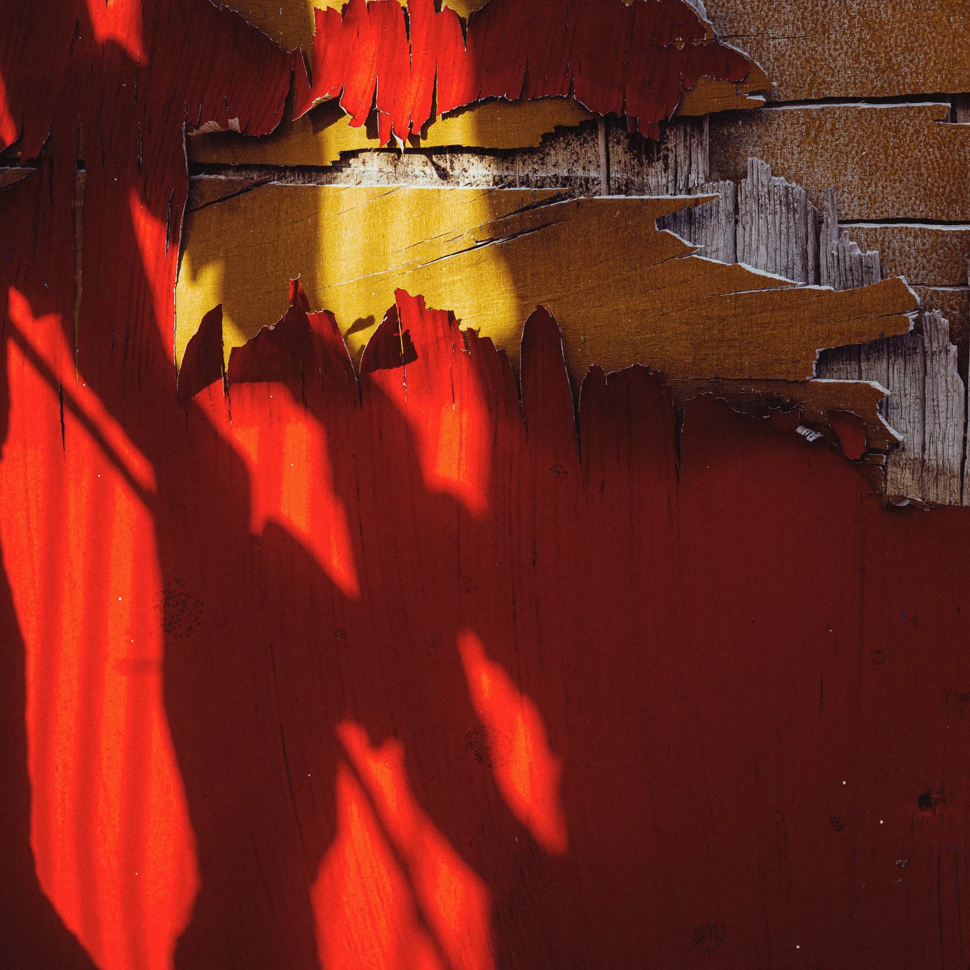 Close-up of distressed, peeling red and yellow paint on a wooden surface, with shadows cast across the weathered wood.