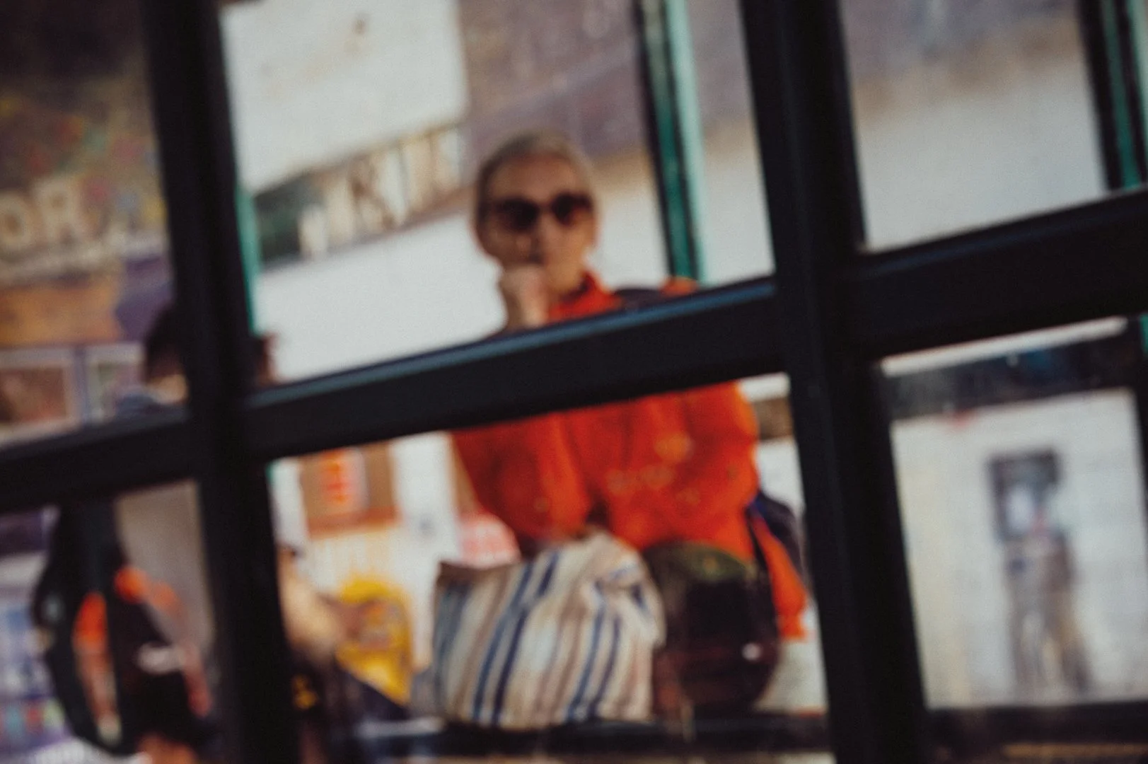 A blurred woman wearing sunglasses and a red shirt, standing behind a window with black grid frames, possibly holding a shopping bag.