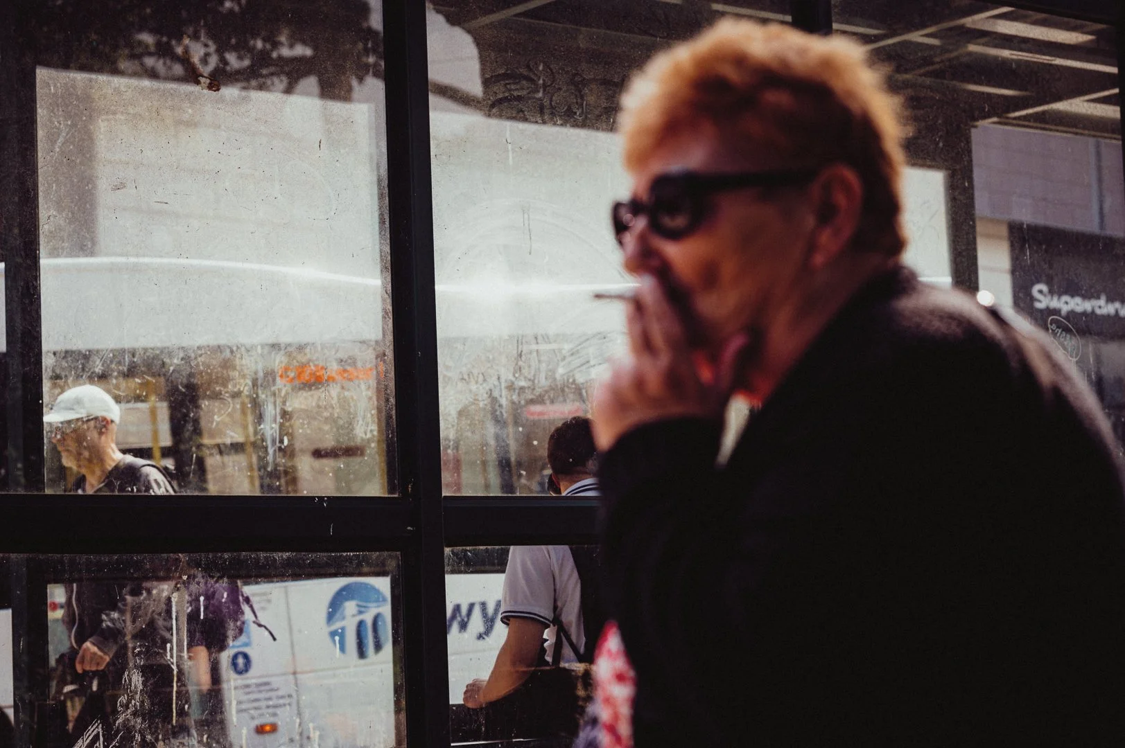 A blurred man with light hair, glasses, and a dark jacket, standing outside at a bus stop or transit station, with other people visible behind an uneven glass window.