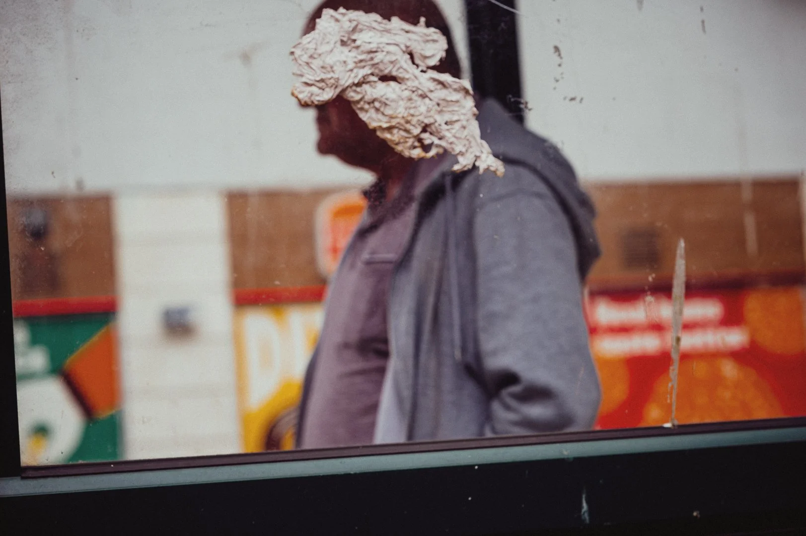 A person wearing a large, textured hat made of a light-colored, crumpled material, standing outdoors with a blurred background featuring colorful signage and a wooden surface.