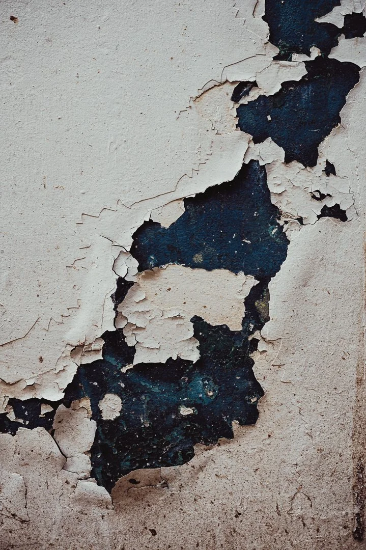 Close-up photograph of peeling white paint on a wall, revealing dark blue and black layers underneath.