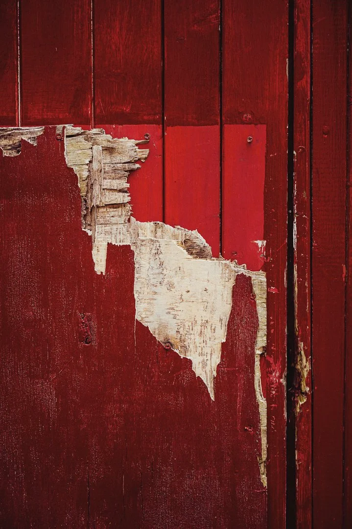 Close-up of a red wooden wall with peeling paint revealing old white paint underneath.