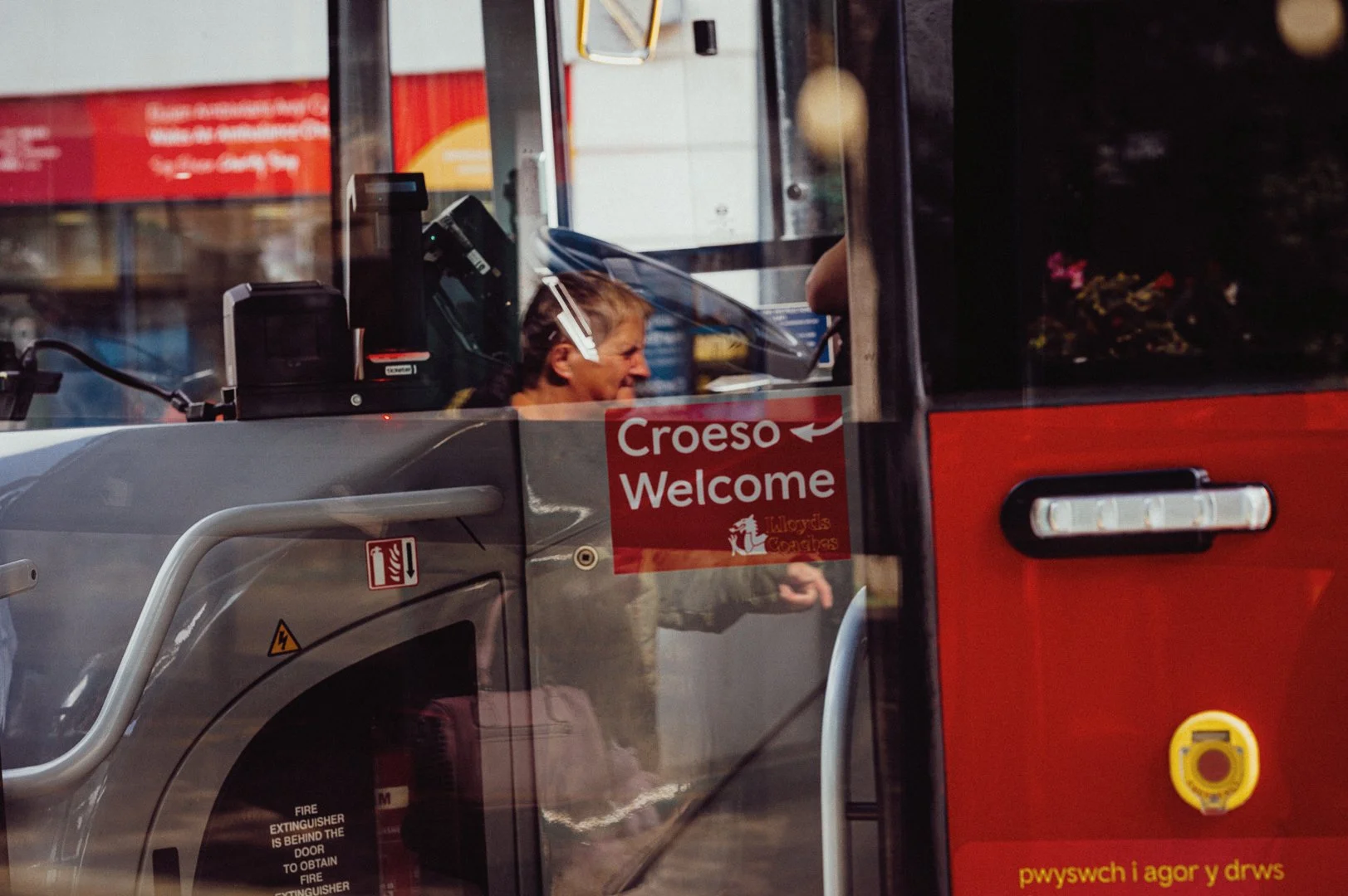 A bus driver seen through a window, with a red sign that says 'Croeso Welcome' and has a Welsh language phrase at the bottom. The driver has short blonde hair and is smiling.
