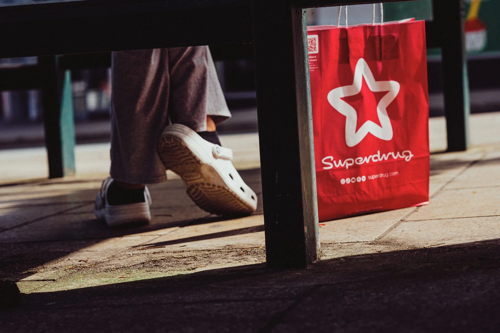 Person's legs and feet wearing sneakers and gray pants, sitting at a table with a red shopping bag labeled Superdrug on the ground beside them.