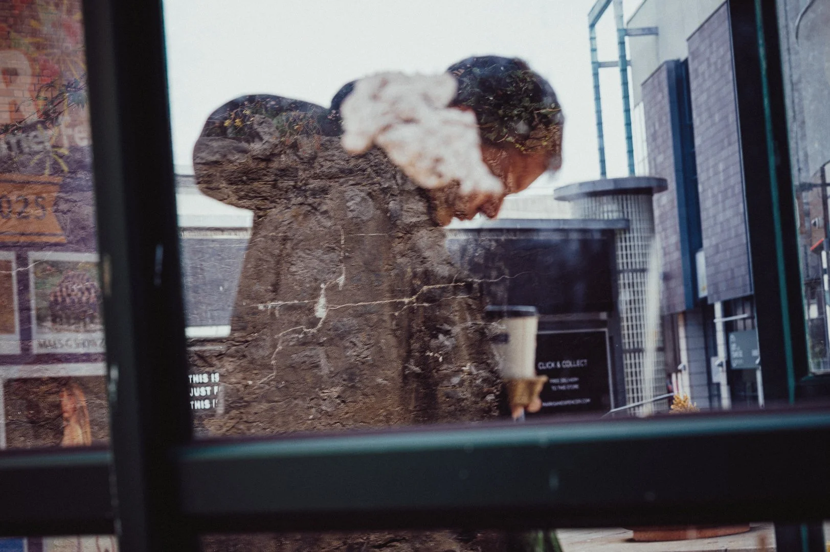 Reflection of a woman with short dark hair, looking down at her coffee cup, seen through a window.