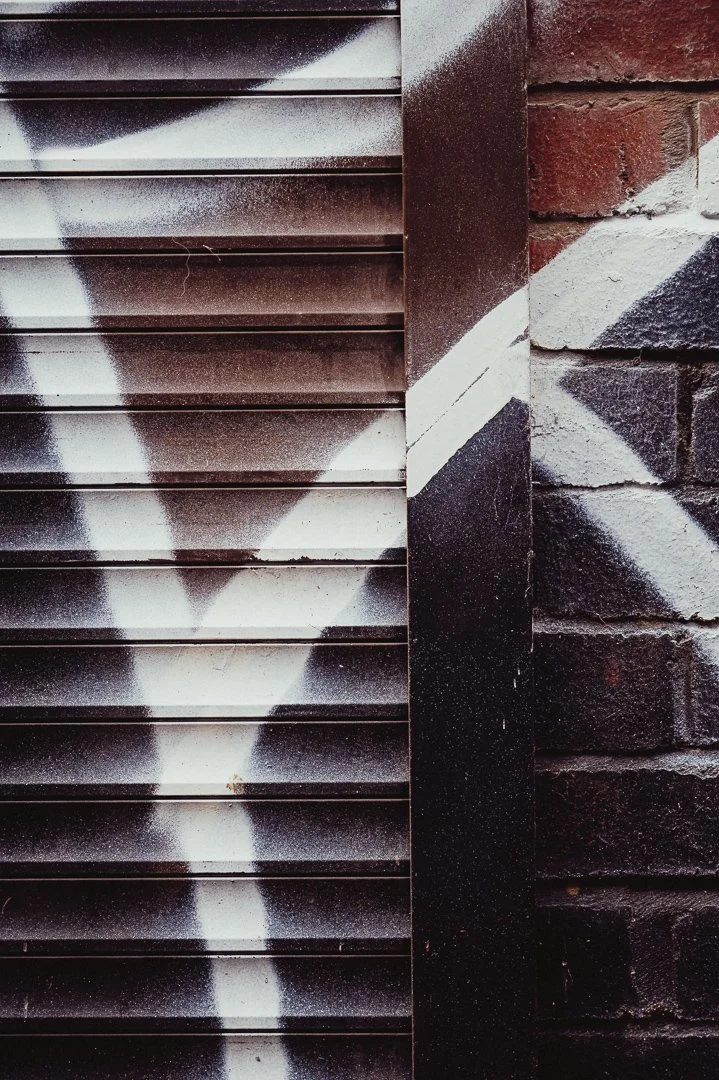 Close-up of a metal vent with graffiti on the slats, next to a brick wall with geometric painted design.