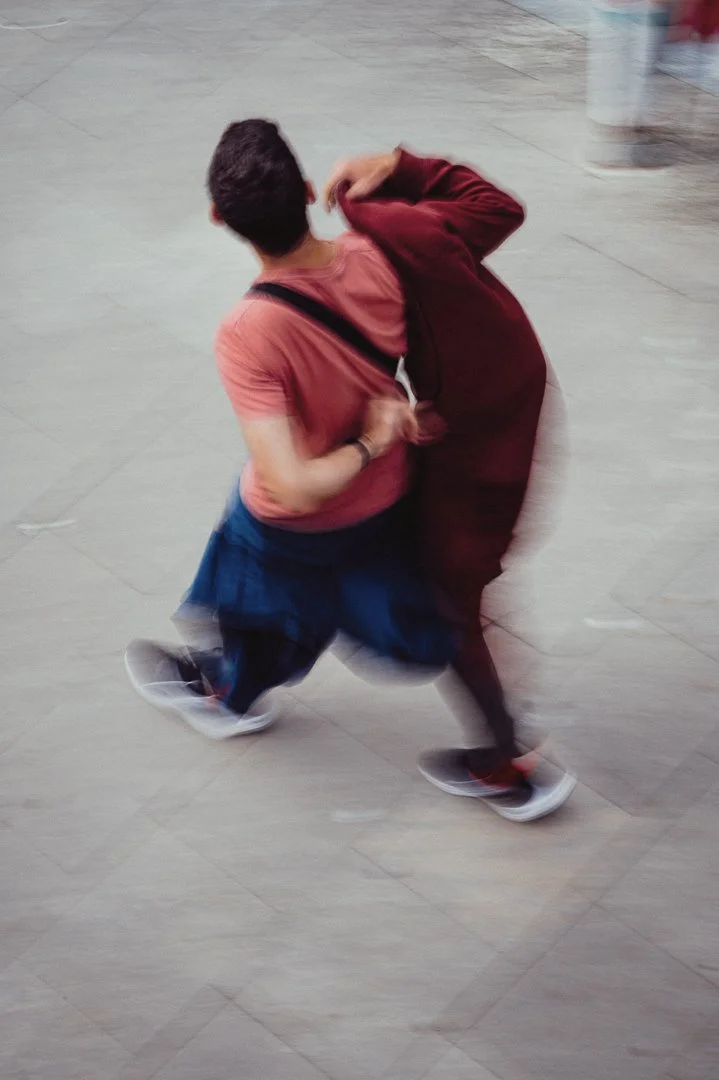 A person in motion captured mid-step on a tiled floor, wearing a pink t-shirt, blue jeans, and sneakers, with a red hoodie or jacket draped over the shoulders.