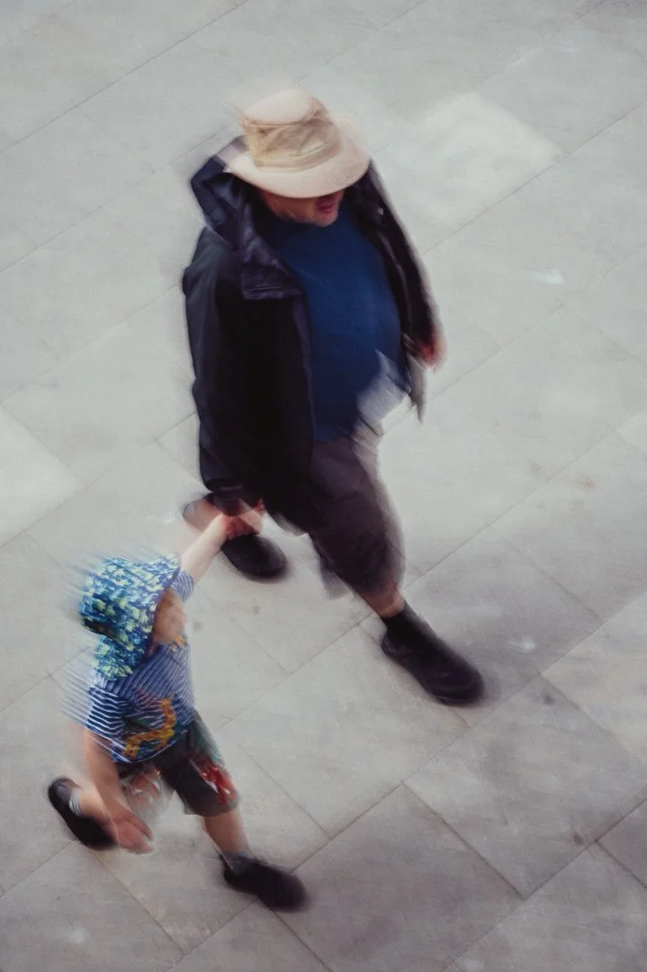 An adult holding a child's hand as they walk on a tiled floor. The adult wears a large sunhat, dark jacket, blue shirt, and shorts. The child wears a colorful hat, striped shirt, and shorts.