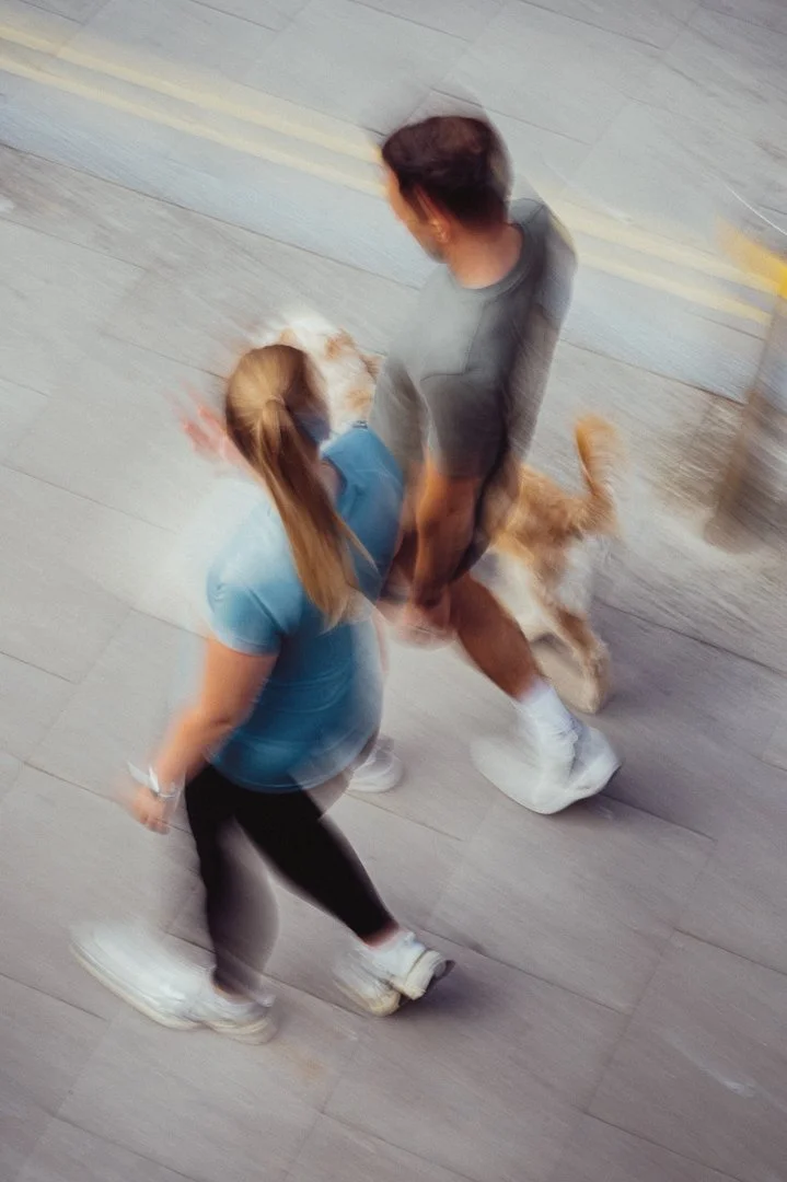A woman with long red hair and a man with short dark hair walking with a golden retriever on a wooden floor.