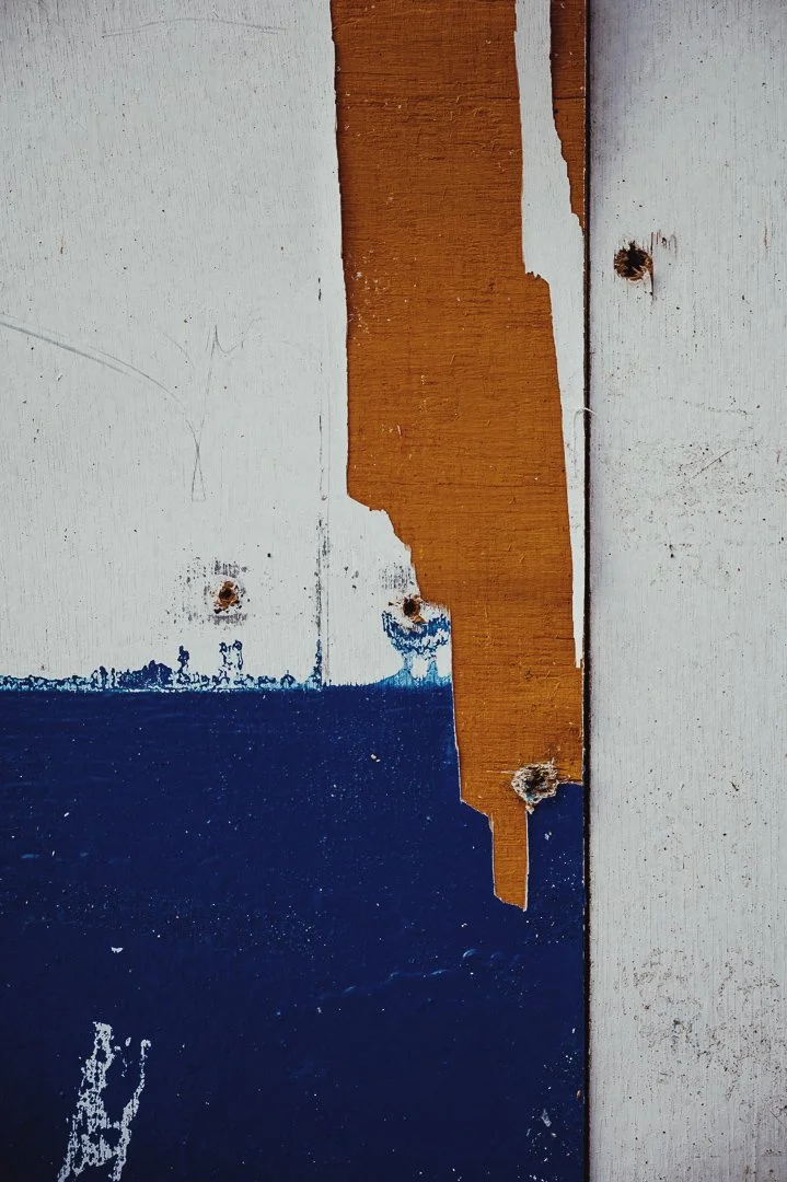 Close-up of peeling paint on wooden surface with white, blue, and orange colors, revealing aged wood underneath.