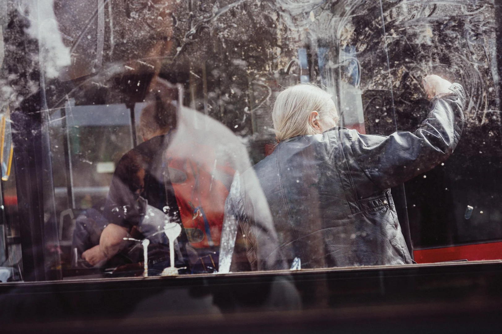 A person with blonde hair in a leather jacket wiping a window of a bus, with another person visible inside the bus and reflections of trees and surroundings on the window.
