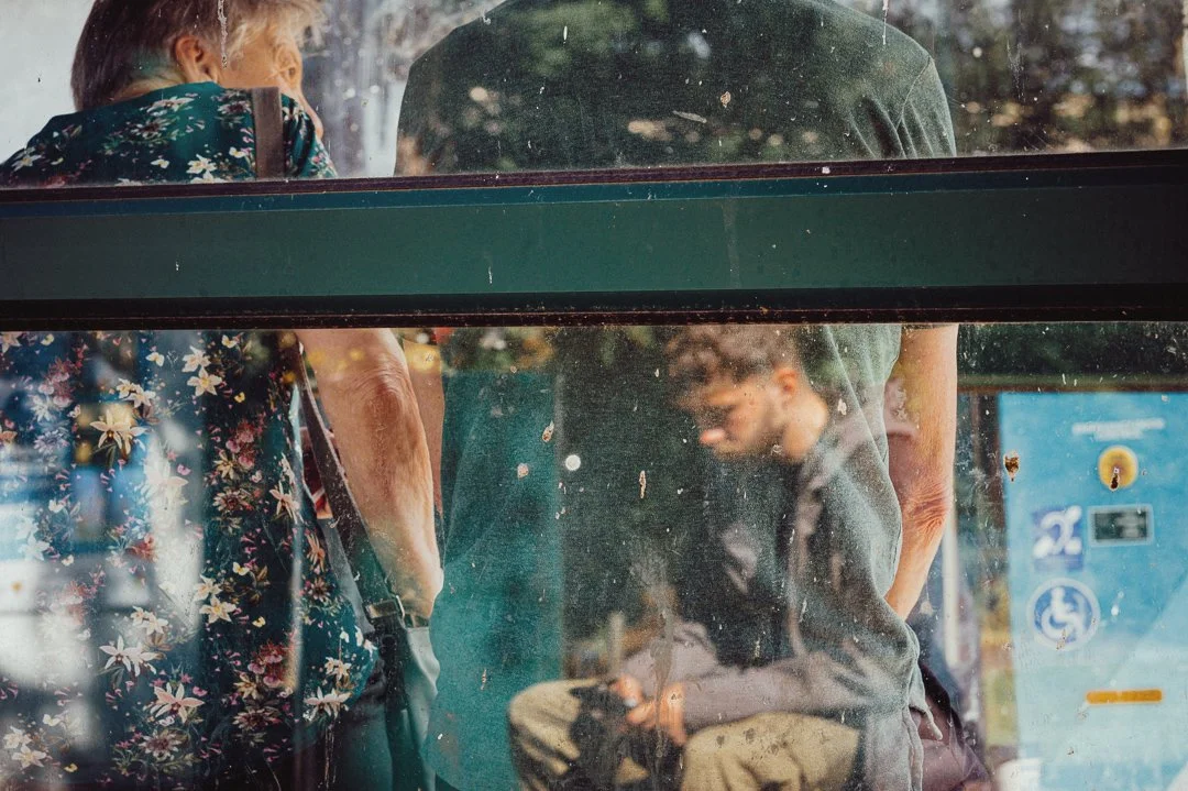 Three people standing outside near a glass window, with smudges and dirt on the glass. Two are adult, one with floral clothing and one with a green shirt, and the third is a young male, sitting and looking down at his mobile device.