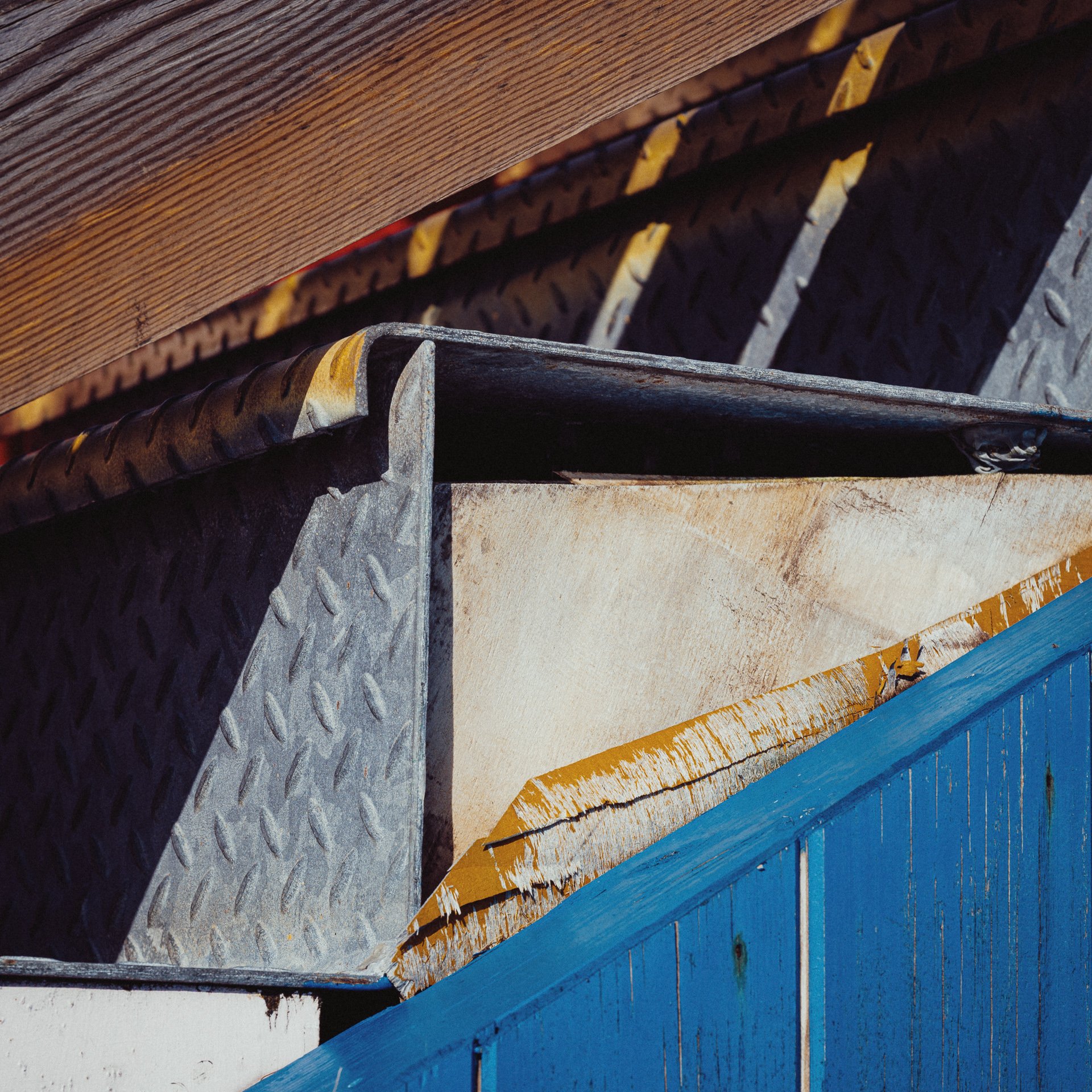 Close-up of a metal container with a diamond plate pattern, partially covered by a weathered yellow plywood sheet and framed by a blue wooden wall, with shadows cast on the scene.