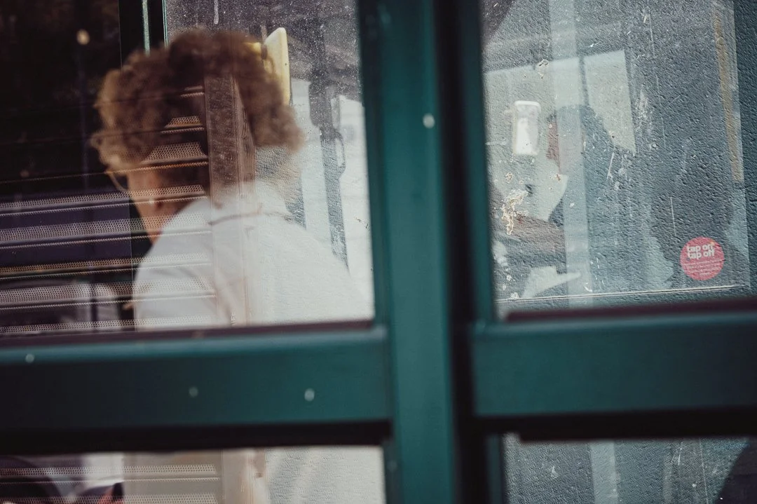 A person with curly hair wearing a white shirt standing inside a building, viewed through a glass window or door with reflections and dirt on the surface.