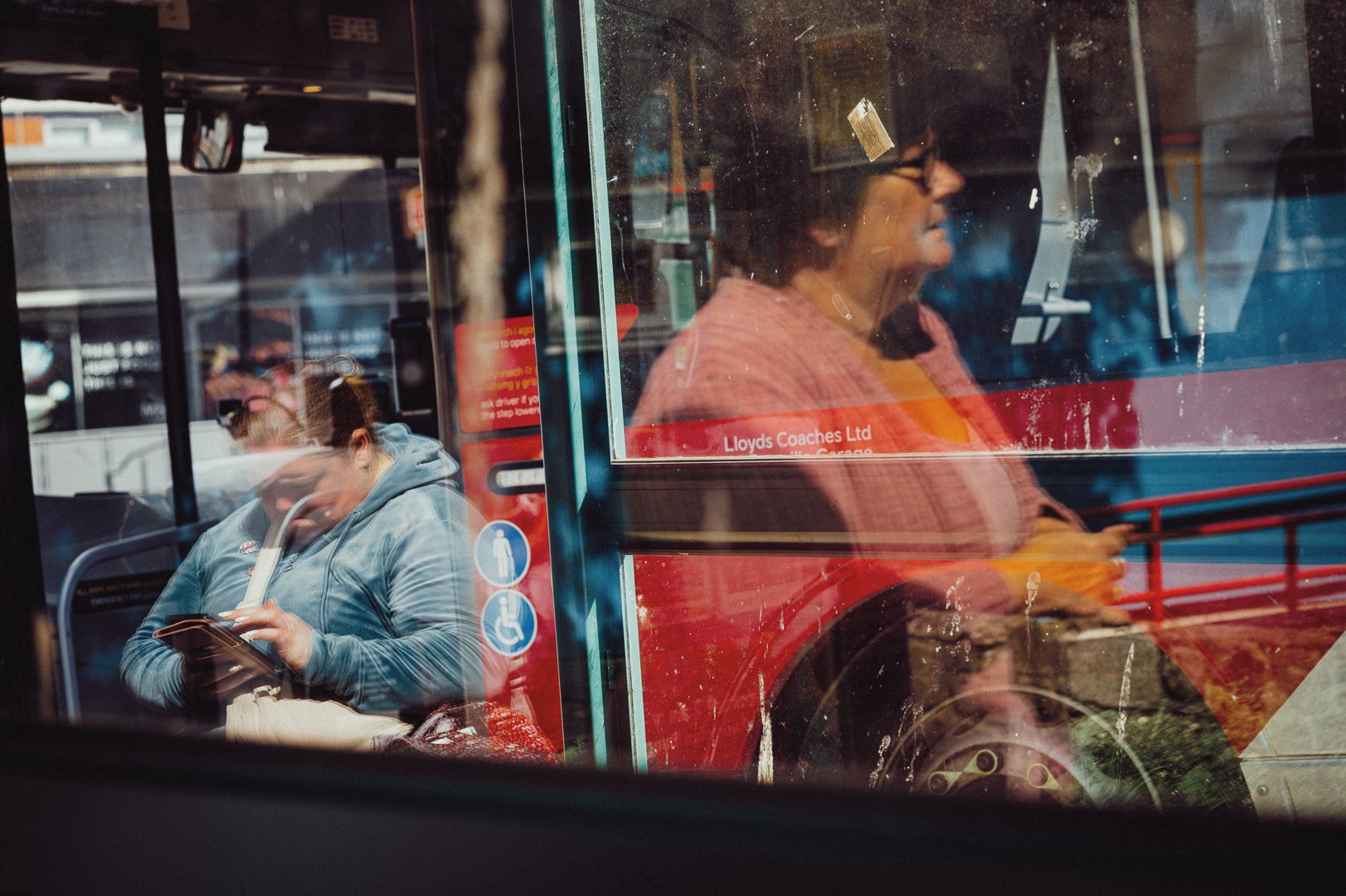 Two women seated on a bus, one in the foreground wearing a blue hoodie and using a smartphone, the other in the background with glasses, wearing a pink sweater, looking outside the window.
