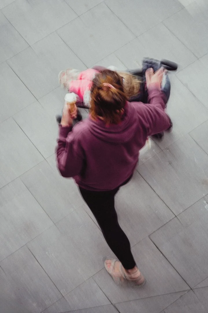 A woman in a purple hoodie and black leggings pushing a young girl in a pink jacket on a shopping cart through a tiled floor.