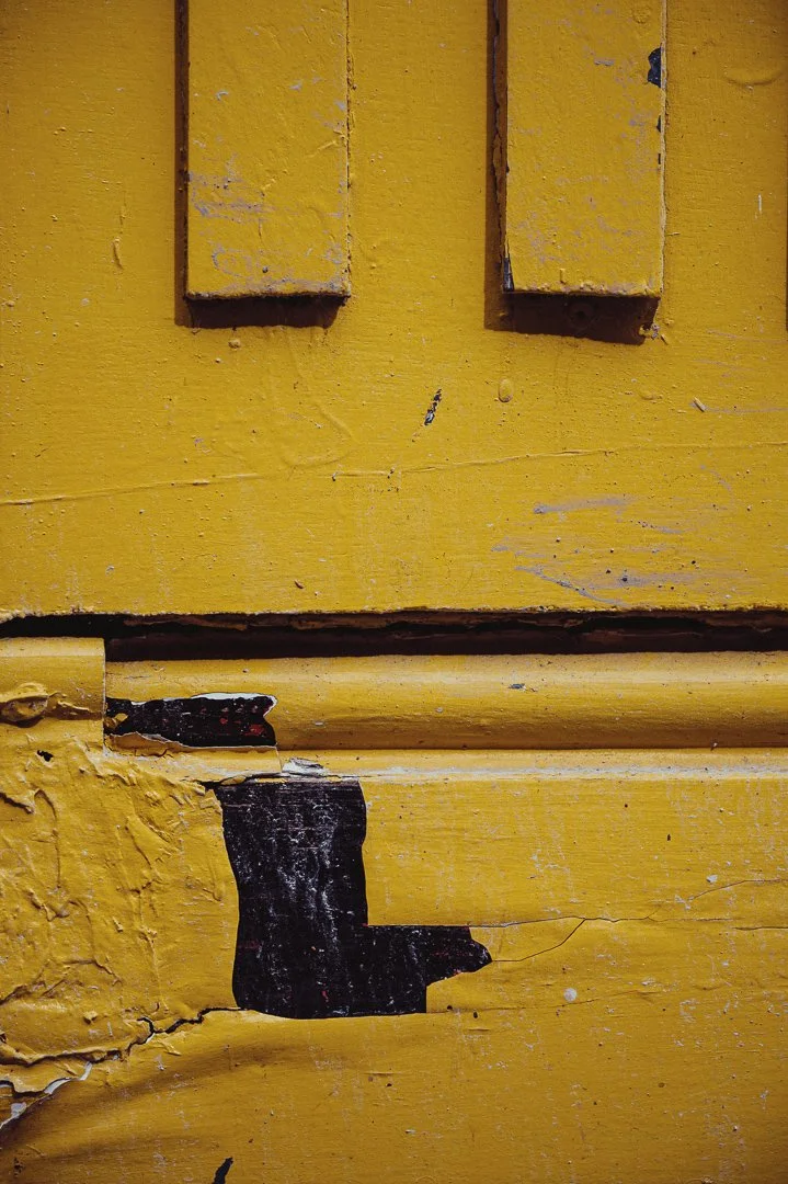 Close-up of a yellow painted wooden surface with cracked and peeling paint, showing layers of paint and wood underneath, and a part of a black painted area.