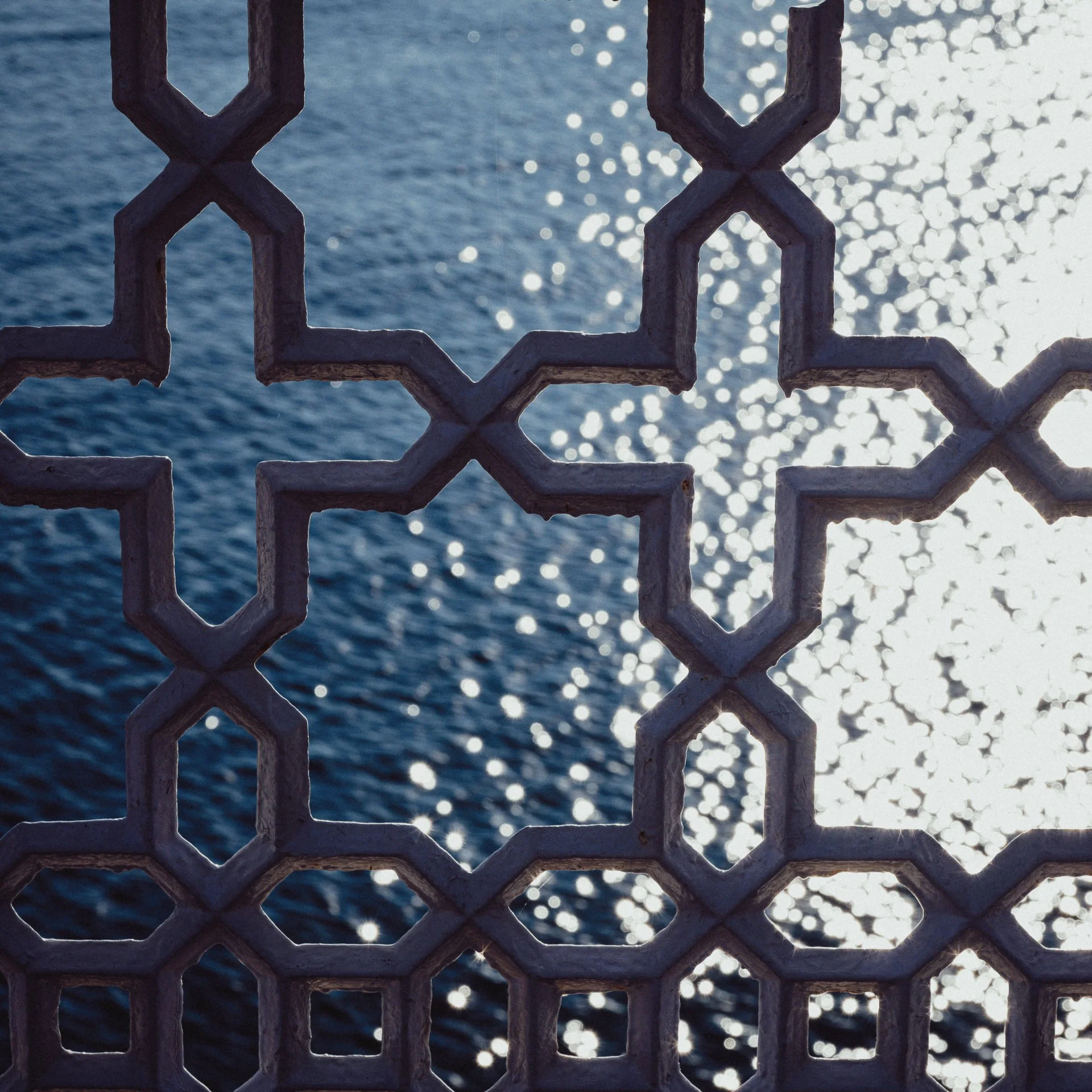 View of water with sunlight reflecting off the surface, seen through a decorative metal railing with geometric patterns.