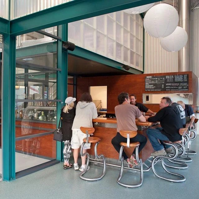 People sitting and standing at a wooden bar counter inside a modern cafe, with a black menu board and white paper lanterns hanging from the ceiling.