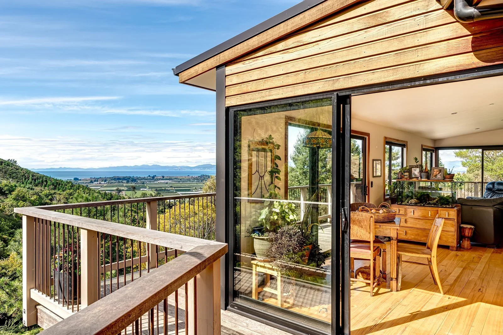 View from a house balcony overlooking a green landscape with mountains and water in the distance, and an open sliding glass door leading inside a room with wooden furniture and houseplants.