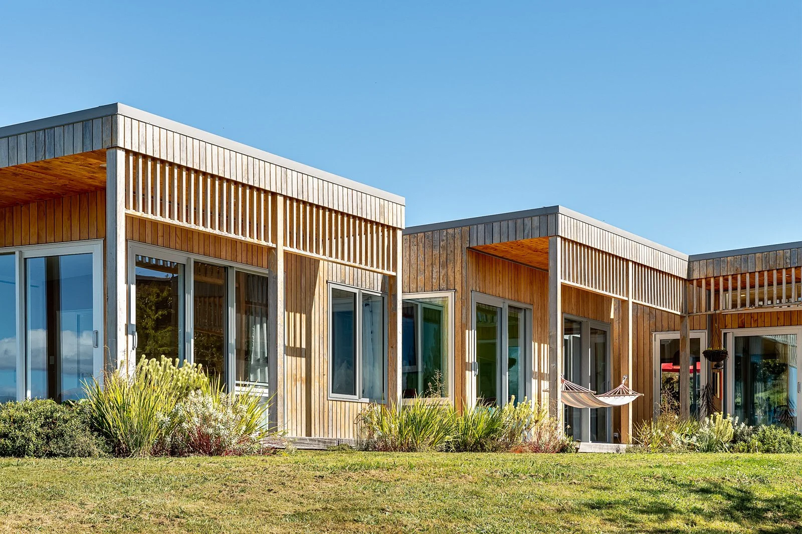 Modern wooden house with large glass sliding doors, surrounded by landscaping and a hammock, under a clear blue sky.