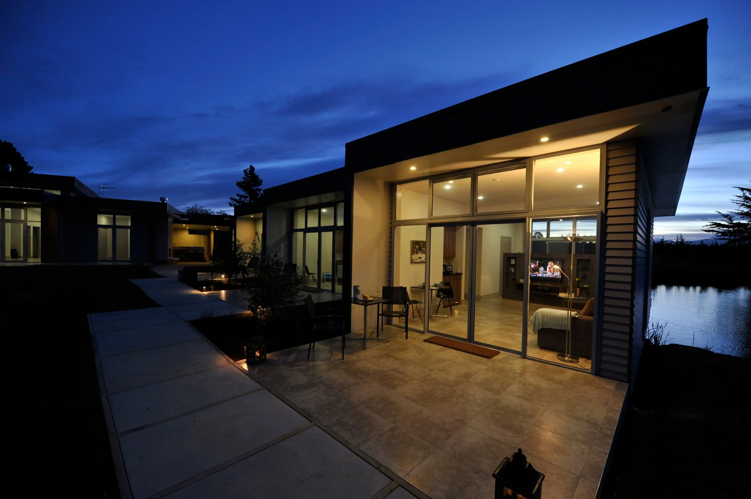 Modern house with large glass windows illuminated from within during twilight, overlooking water with a clear blue sky overhead.