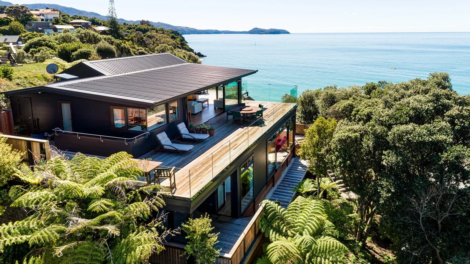 Modern house with large balcony overlooking the ocean, surrounded by trees and greenery.
