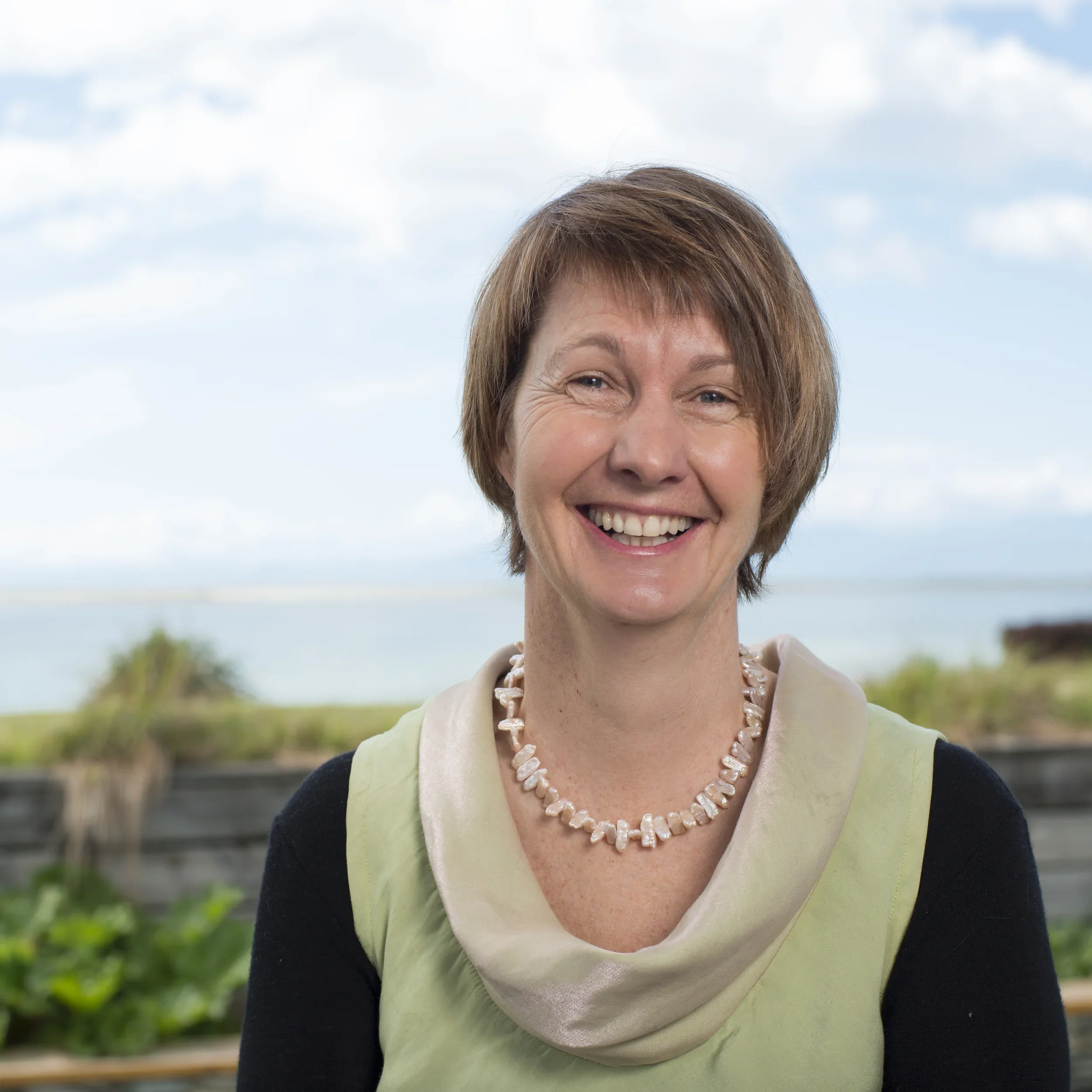 Gabrielle Bell, smiling middle-aged woman with short brown hair, wearing a black top and a light green scarf, outdoor setting with a body of water and a cloudy sky in the background.