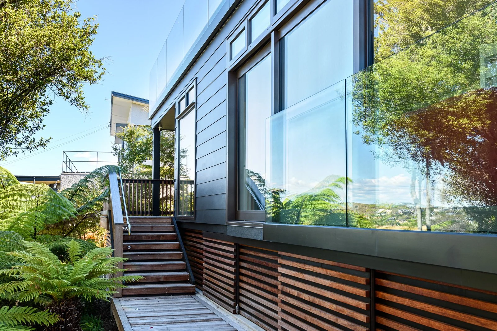Exterior view of a modern house with dark siding, large glass windows, wooden stairs, a glass railing, and lush greenery in a sunny landscape.