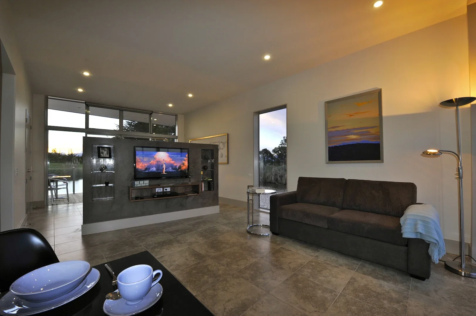 Living room with a brown sofa, wall art, a television on a gray partition, and a view of water outside through glass doors.