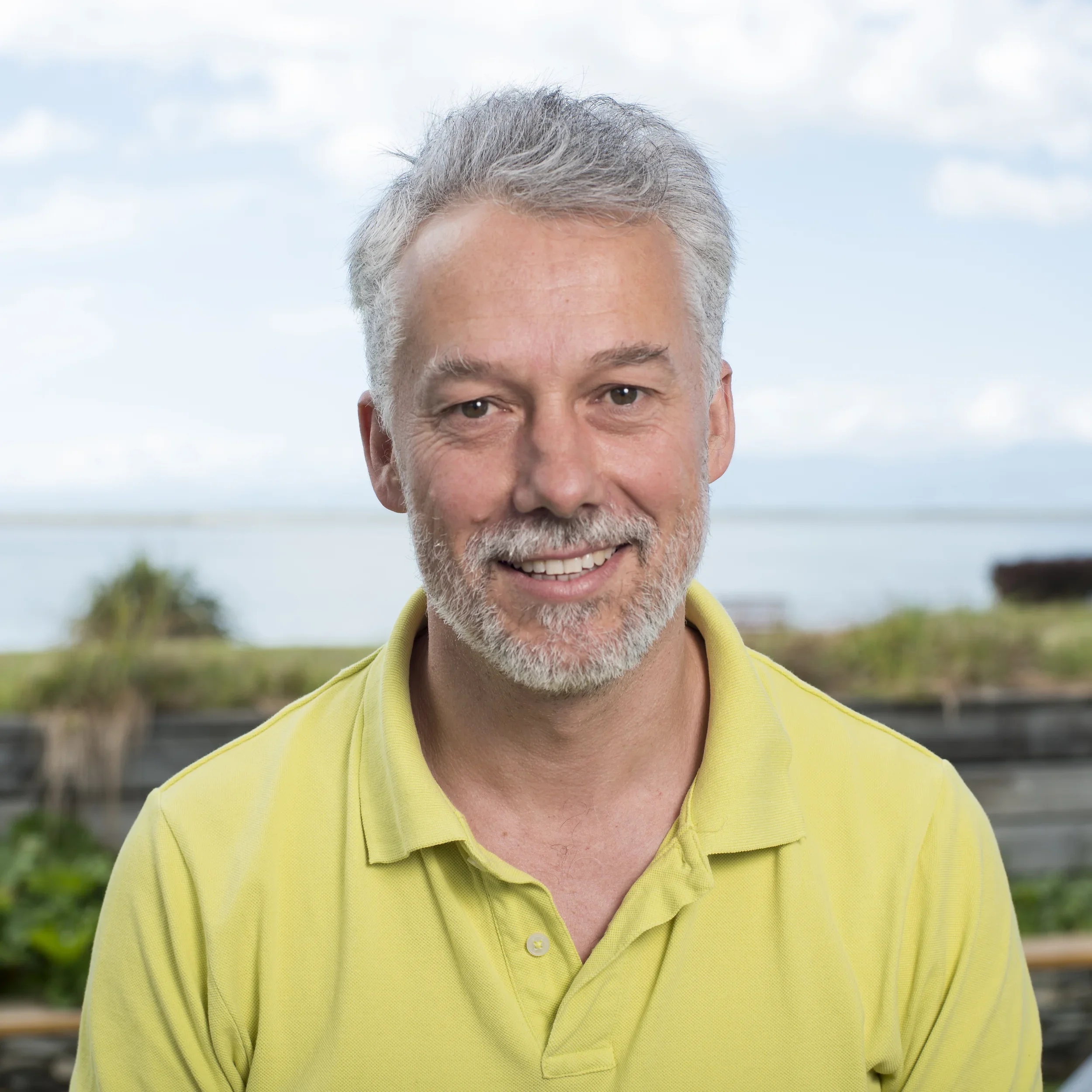 Andrew Stephenson - smiling man with gray hair and beard wearing a yellow polo shirt outdoors near water and greenery.