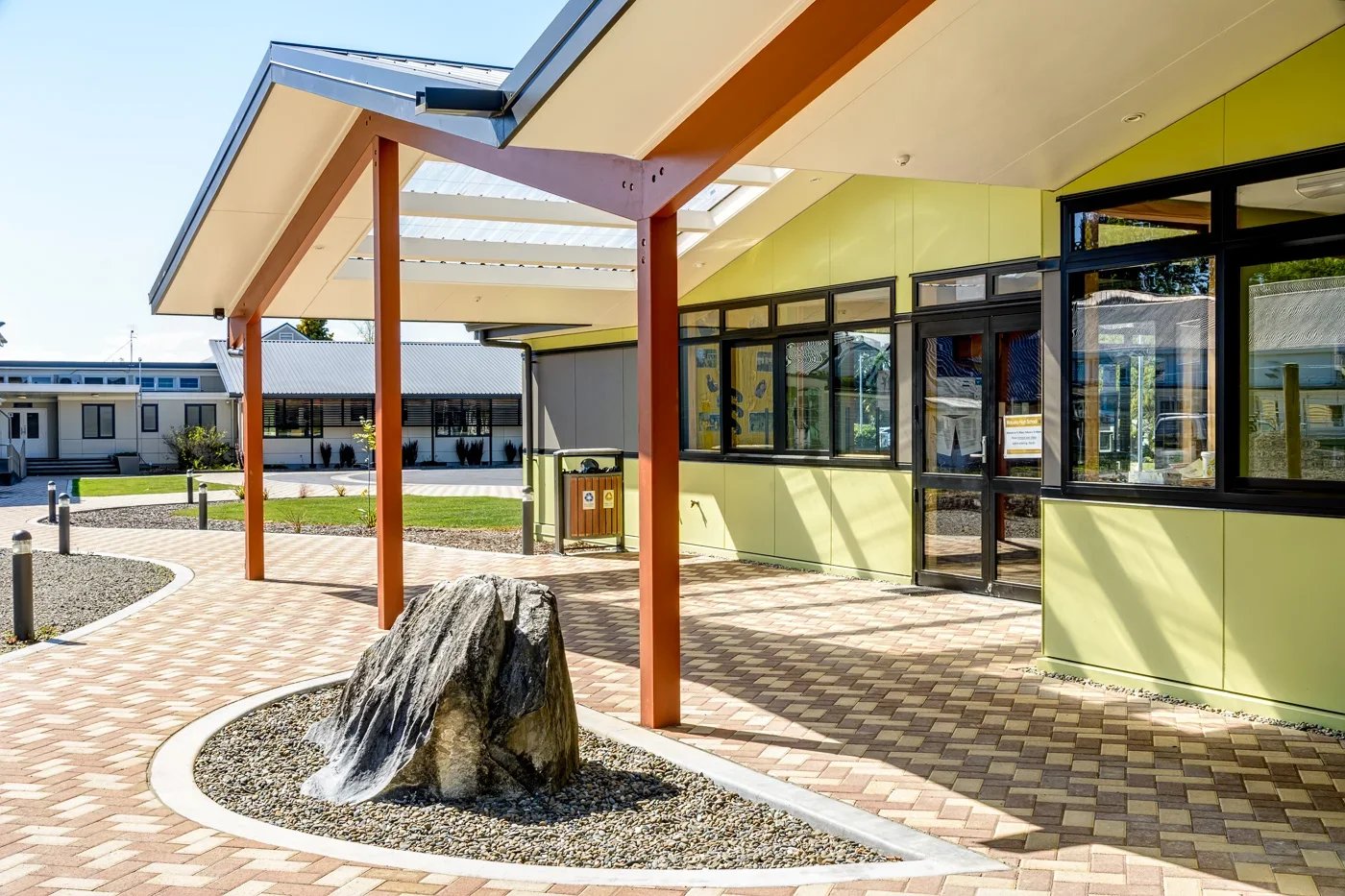 Exterior of a modern building with large windows, a covered walkway with support beams, a large rock, and landscaped surroundings.