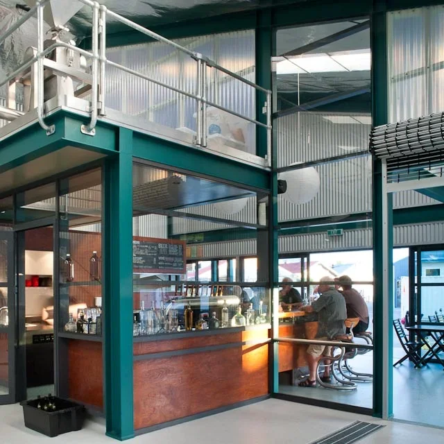 Interior of a modern cafe or coffee shop with a bar counter, some customers sitting at the bar, and large windows letting in natural light.