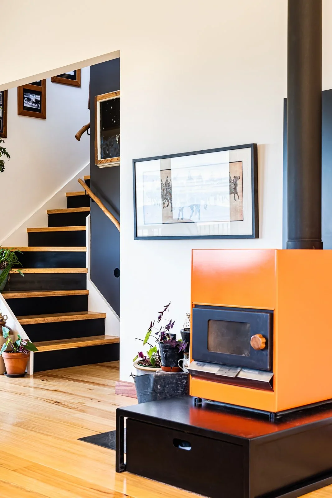 Interior of a home with wooden stairs, framed artwork on the wall, potted plants, and a bright orange wood stove on a black table.