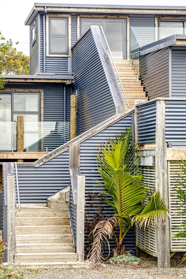 A multi-level building with gray corrugated metal siding, outdoor stairs, and a wooden deck, featuring large windows and tropical plants in the foreground.