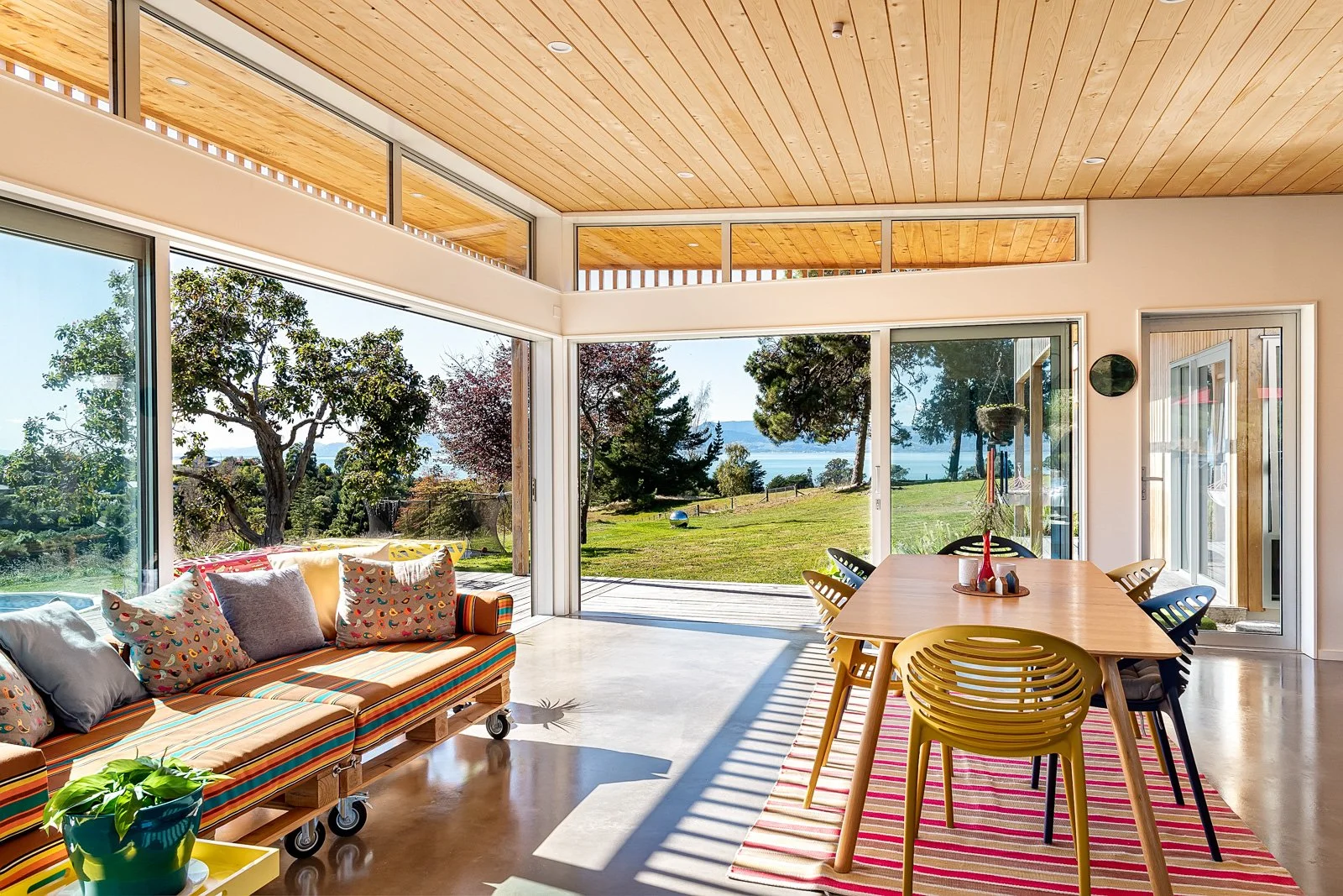 Bright living room with large glass sliding doors opening to a grassy outdoor area with trees and a body of water in the distance. The room has a wooden ceiling, white walls, and colorful furniture including a striped couch with cushions and a wooden dining table with yellow chairs. A pink and white striped rug is under the dining table.