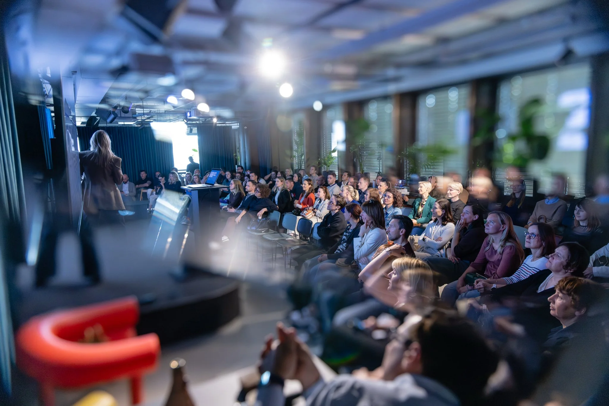 A woman giving a presentation to a large audience in a conference room. The audience appears attentive, and the room is well-lit with natural light coming through large windows.