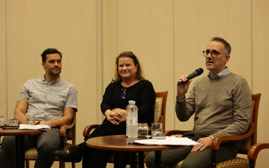 Three seated participants in discussion; the participant on the right holds a microphone.