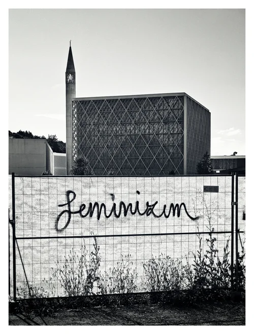 A modern building and tall spire behind a fence marked with graffiti, photographed in black and white