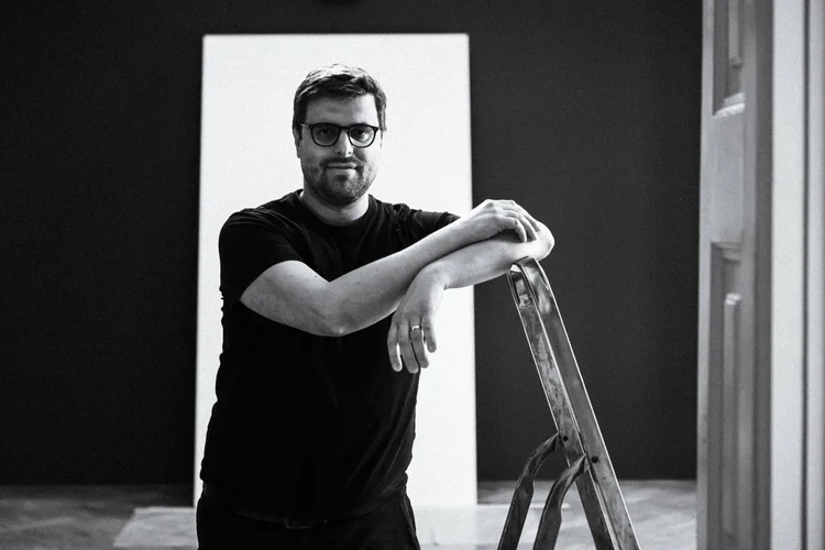 A man with glasses rests his folded arms on a ladder, photographed in black and white