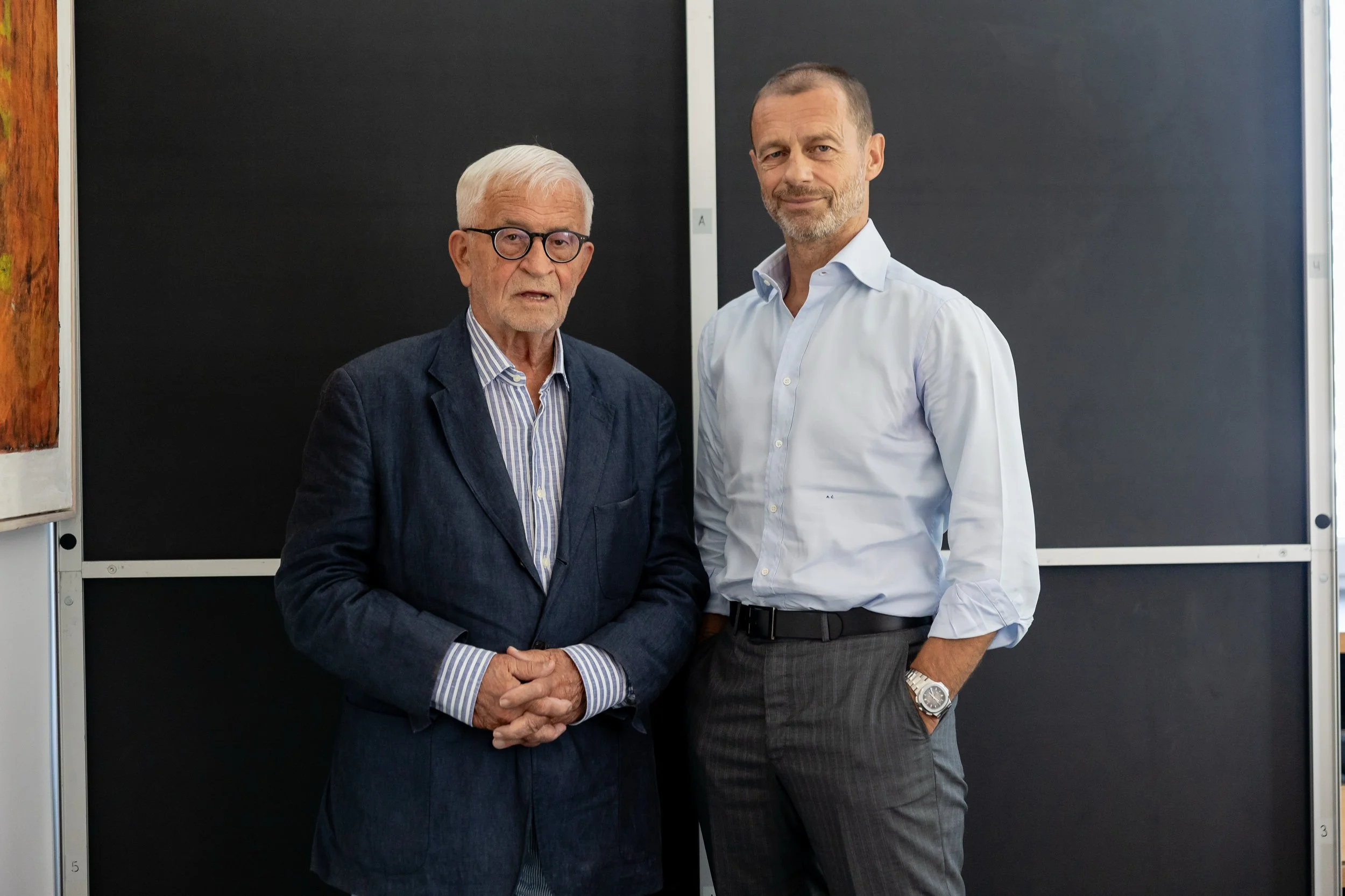 Two men standing side by side in front of a blackboard. The older man on the left has white hair, glasses, and is wearing a dark blazer, striped shirt, and no tie. The younger man on the right has short hair, a beard, and is wearing a white dress shirt with the sleeves rolled up and a wristwatch.
