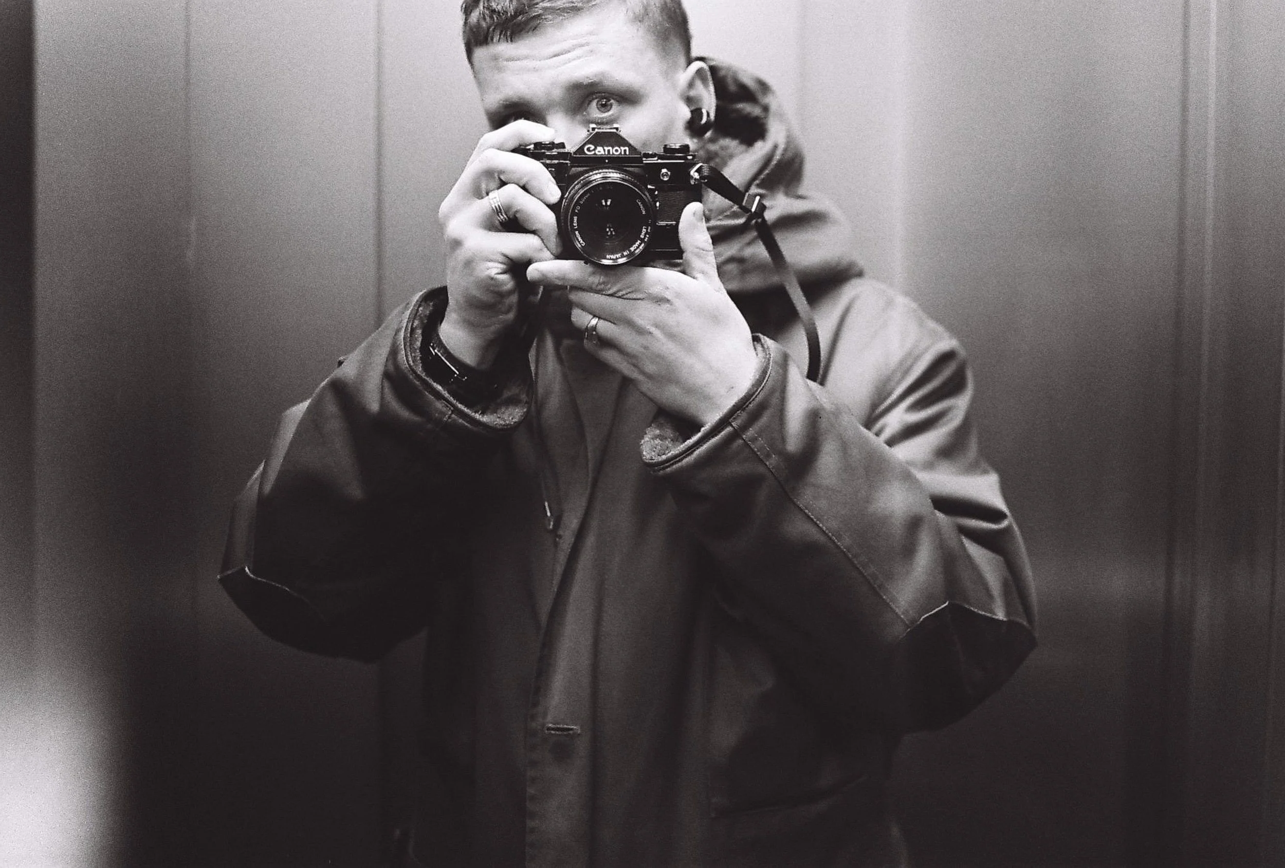 A person with earrings and rings taking a black-and-white photograph in an elevator mirror with a Canon camera.