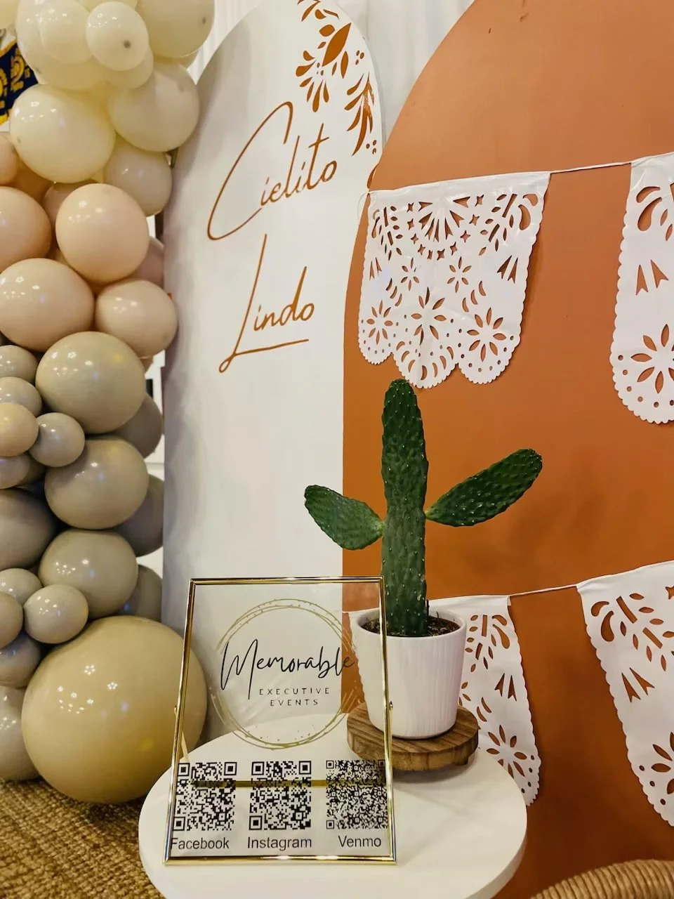 Decorative display featuring a potted cactus on a wooden coaster, a sign that reads 'Memorable Executive Events' with QR codes for Facebook, Instagram, and Venmo, surrounded by beige balloons, a white and orange backdrop with festive cutouts, and a sign with the words 'Cielito Lindo'.