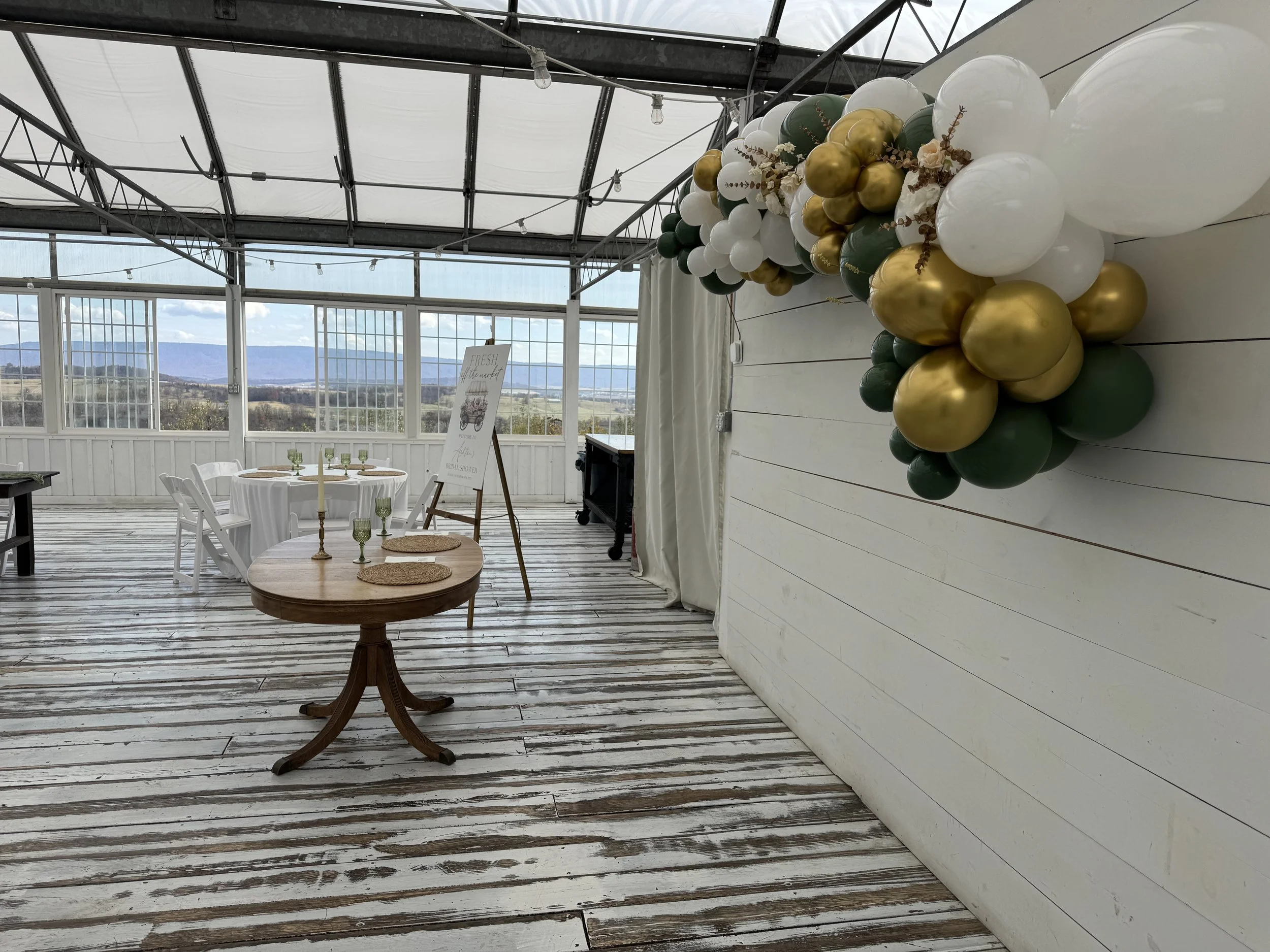 Indoor celebration setup with a balloon garland in white, green, and gold on a white wooden wall, with round tables and chairs, a sign on an easel, and scenic mountains through large windows.