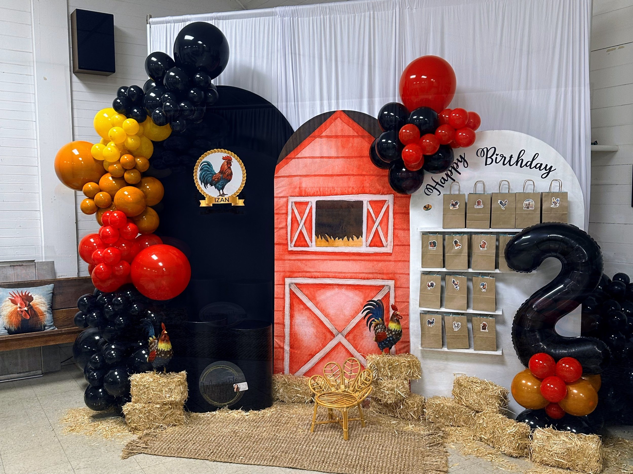 Dessert table decorated with a farm theme including balloons, hay bales, a painted barn backdrop, and goodie bags. The decoration features a rooster motif and a large black number '2' balloon, indicating a second birthday celebration.