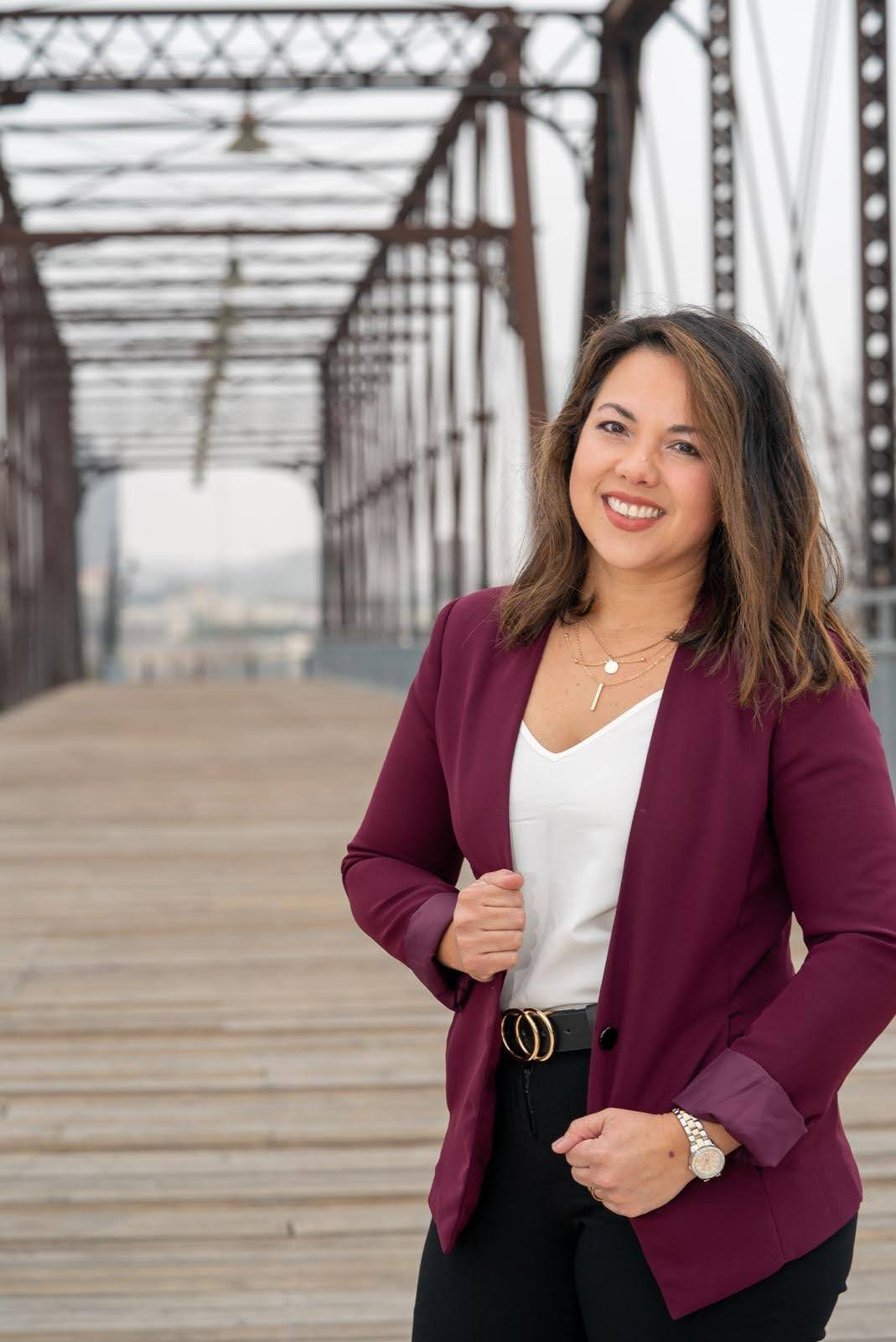 A woman with shoulder-length brown hair, wearing a maroon blazer, white top, black pants, and jewelry, standing on a wooden bridge with metalframework in the background, smiling at the camera.