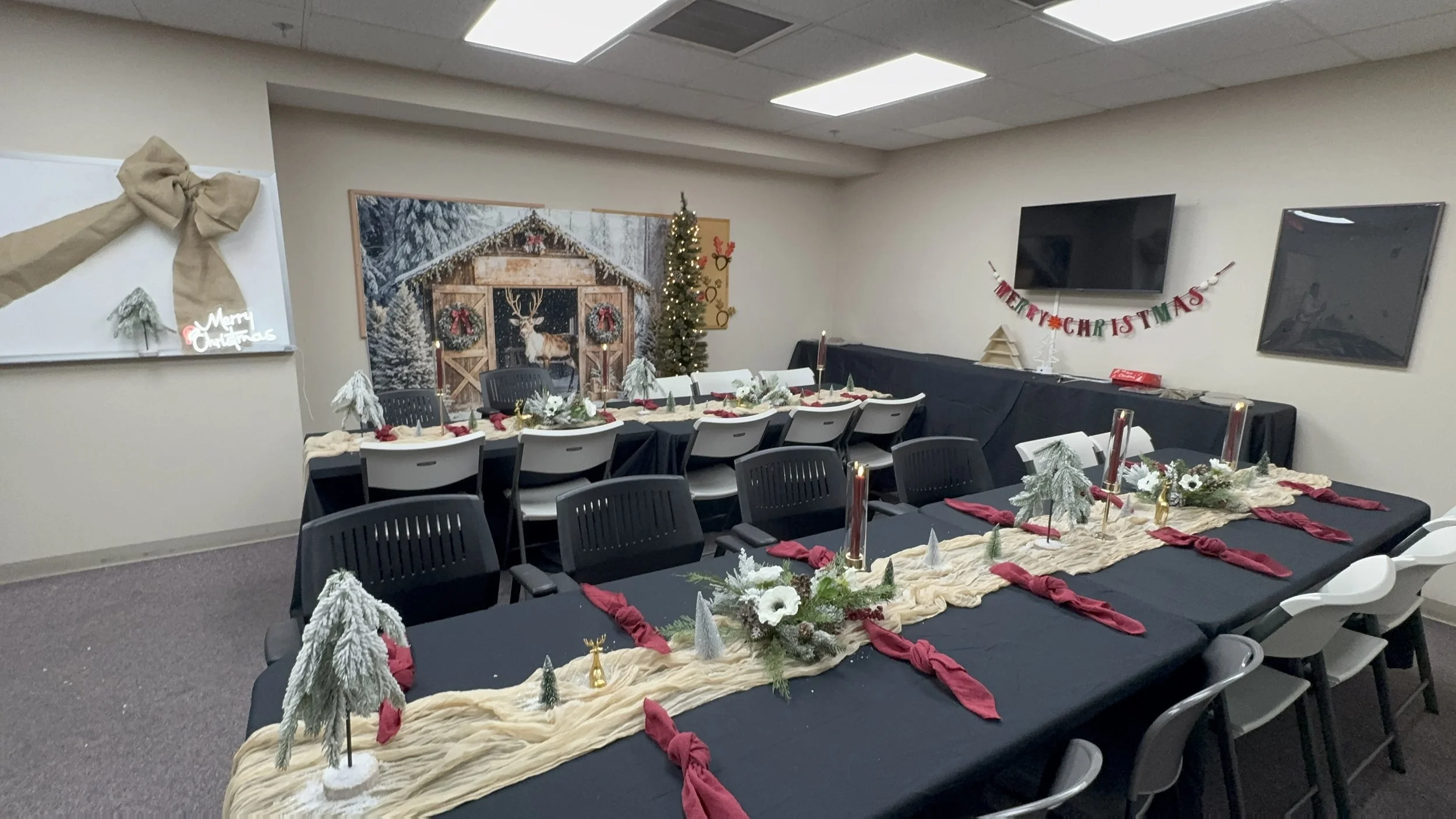 A room decorated for Christmas with long tables covered in black tablecloths, red napkins, and festive centerpieces featuring white flowers, pinecones, and greenery. Small Christmas trees, candles, and reindeer figurines are on the tables. A Christmas tree stands in the corner, and a large winter scene mural of a snowy forest and a wooden cabin with a reindeer and wreaths decorates the wall. A “Merry Christmas” banner hangs on the wall, and a neon “Merry Christmas” sign is on a whiteboard with a burlap bow.
