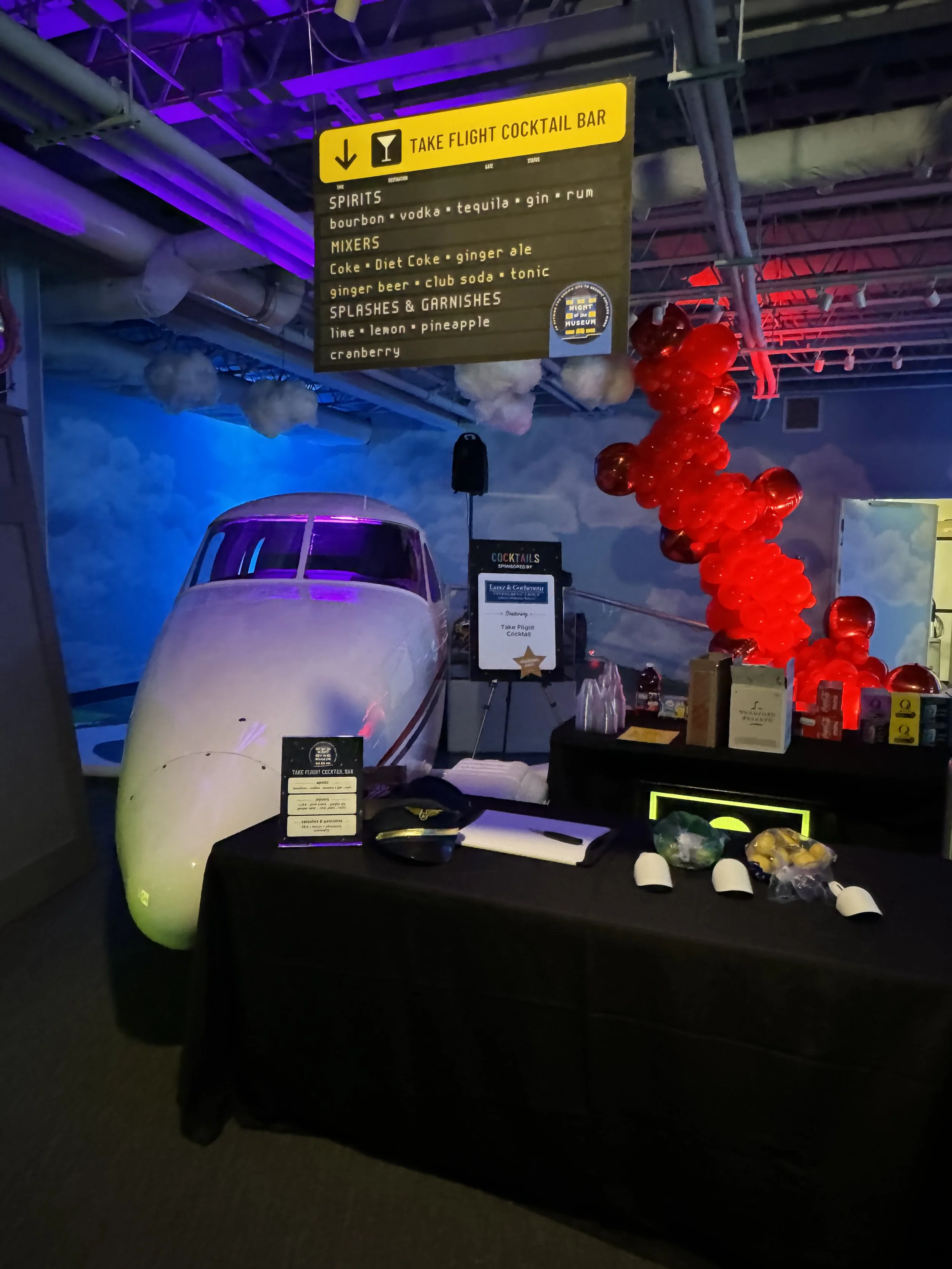 An indoor scene at the flight museum with a retro airplane replica, black table with drink menus, and a colorful balloon decoration in red. There is a large yellow sign hanging from the ceiling with a flight cocktail menu listing spirits, mixers, and garnishes. The background has cloud-themed wall art and purple lighting.